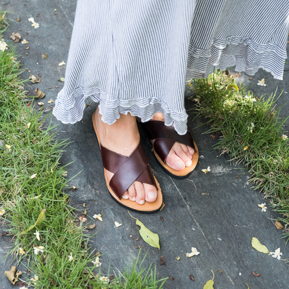 Feet in brown sandals on slate, beneath a striped skirt with ruffled hem, near grass and leaves.
