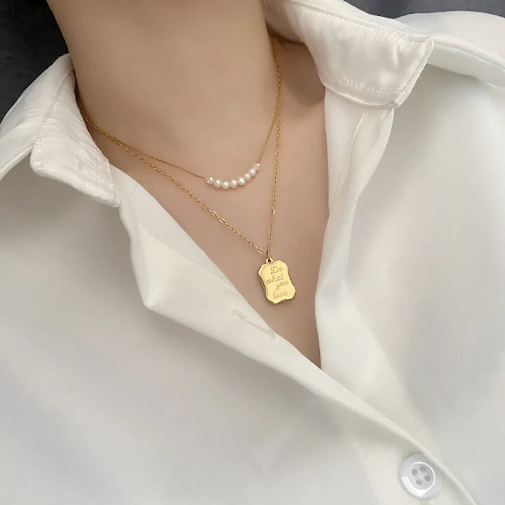 Woman wearing a white shirt with layered necklaces; pearls and a gold pendant with text.