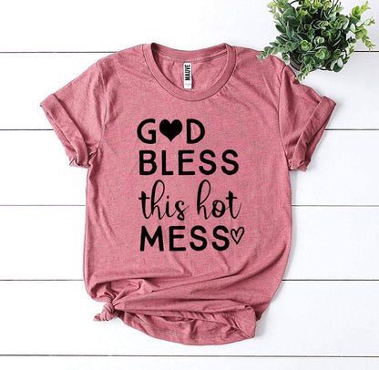 Pink T-shirt with "God Bless This Hot Mess" text and heart icon, placed on white wooden surface.