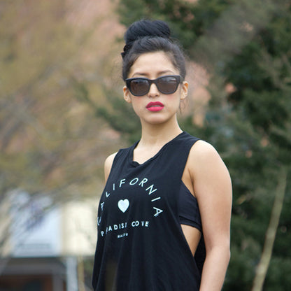 Woman with sunglasses and red lipstick in black "California Paradise Cove" tank top outdoors.