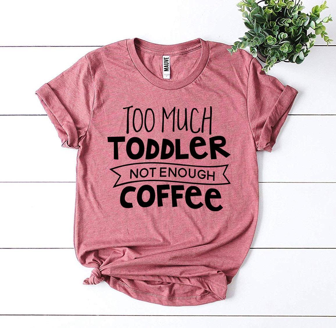 Pink T-shirt with bold black text: "Too Much Toddler Not Enough Coffee," next to a small plant.