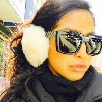 Woman with large sunglasses and white earmuffs in sunlight, wearing a black outfit.