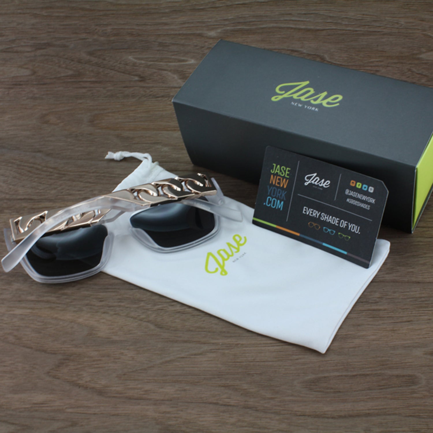 Sunglasses with gold chain arms on a white pouch; black box and card on a wooden surface.