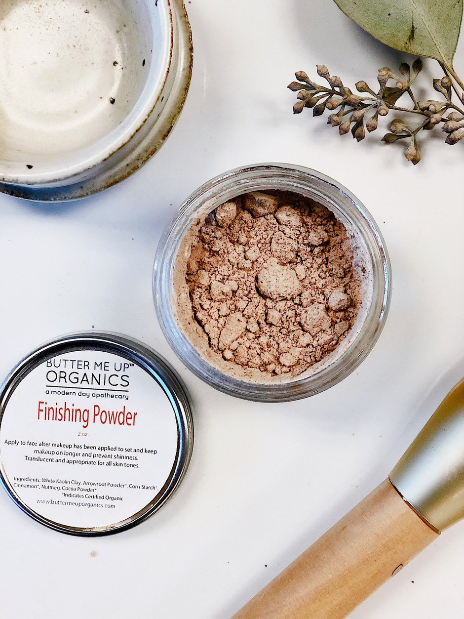 Open jar of finishing powder, labeled lid beside it, brush, ceramic dish, and branch on white background.