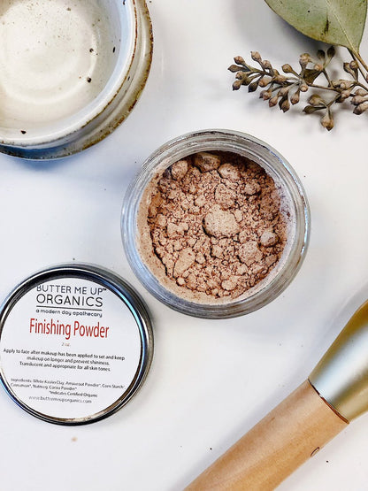 Open jar of finishing powder, labeled lid beside it, brush, ceramic dish, and branch on white background.