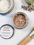 Open jar of finishing powder, labeled lid beside it, brush, ceramic dish, and branch on white background.