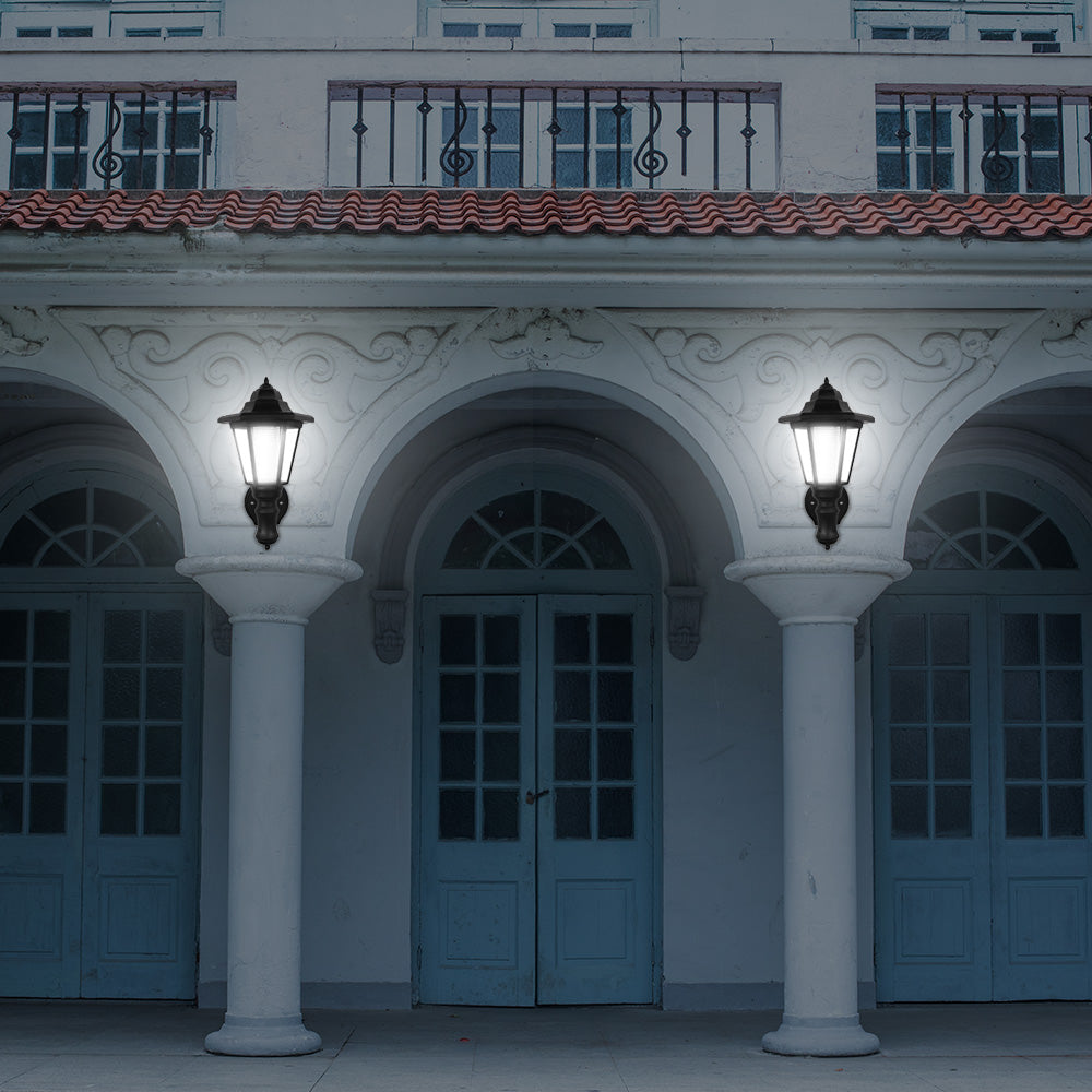 Illuminated porch with two black lanterns on white pillars, arched doors, and wrought iron balcony.