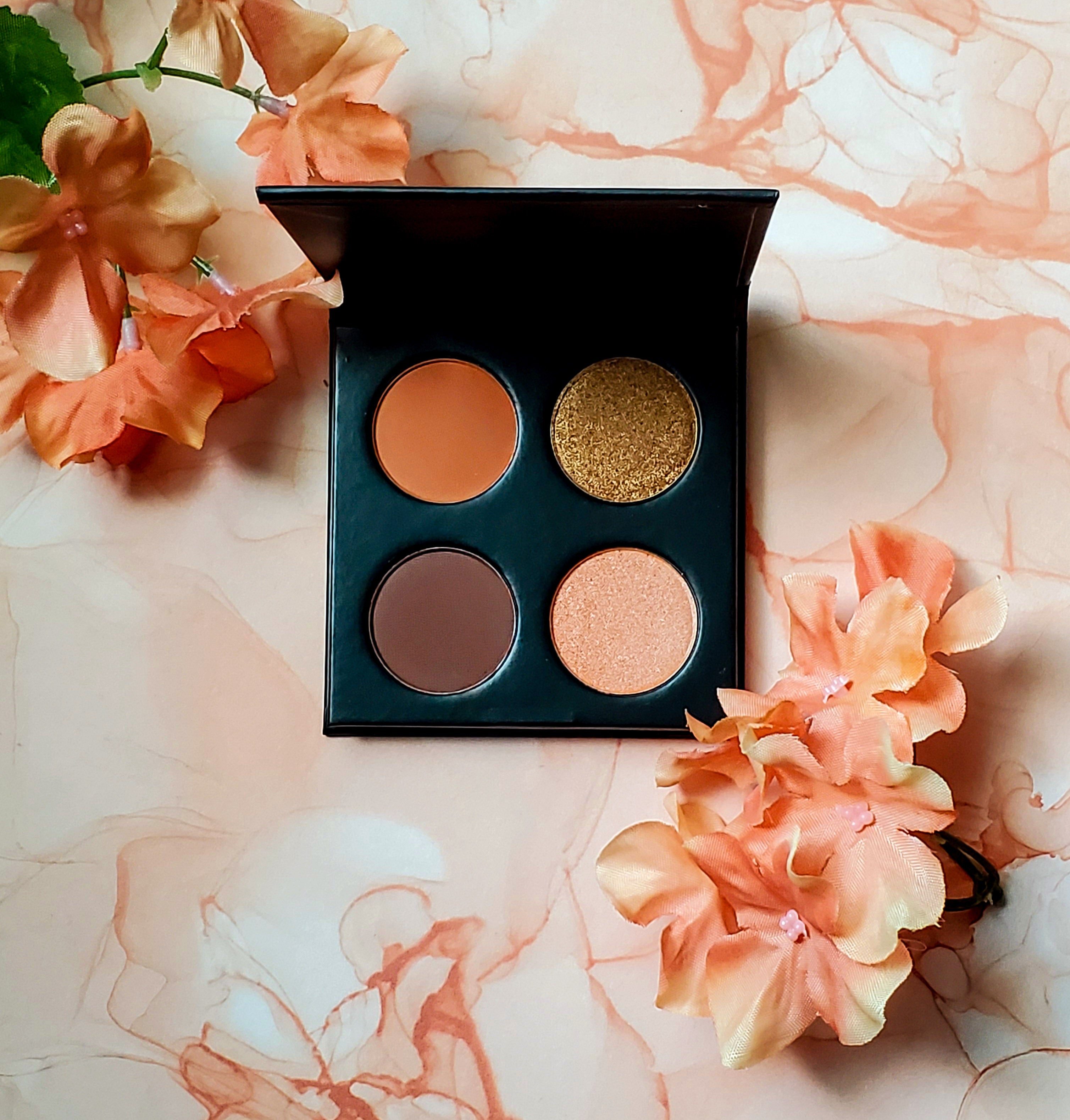 Makeup palette with four warm shades and orange flowers on a peach marbled background.