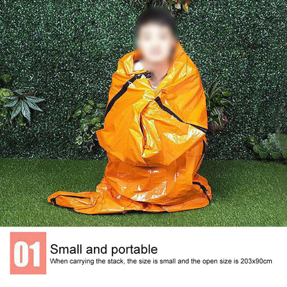 Person wrapped in an orange emergency blanket, against a green leafy background, sitting on grass.