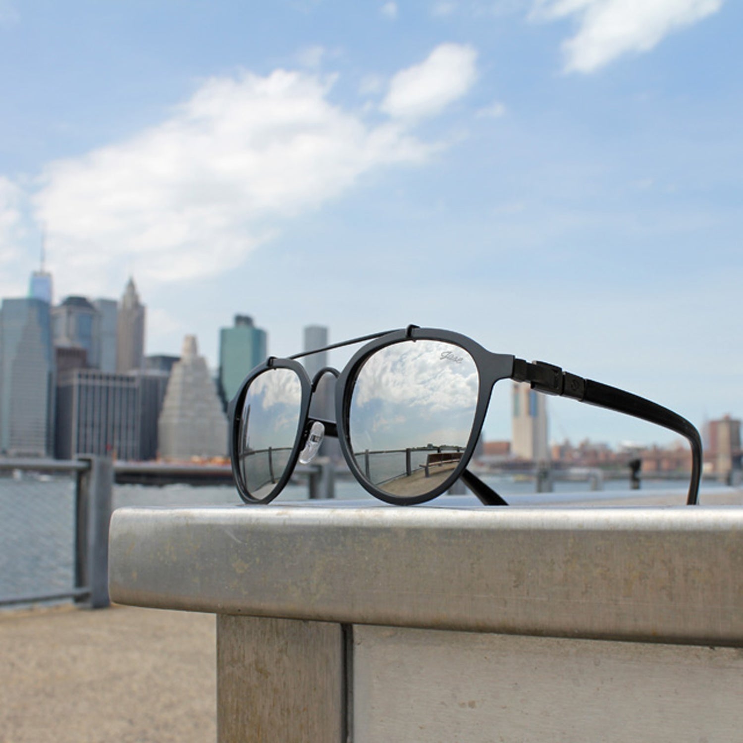 Sunglasses reflecting blue sky and clouds; blurred city skyline and railing in the background.