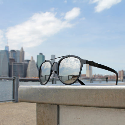 Sunglasses reflecting blue sky and clouds; blurred city skyline and railing in the background.