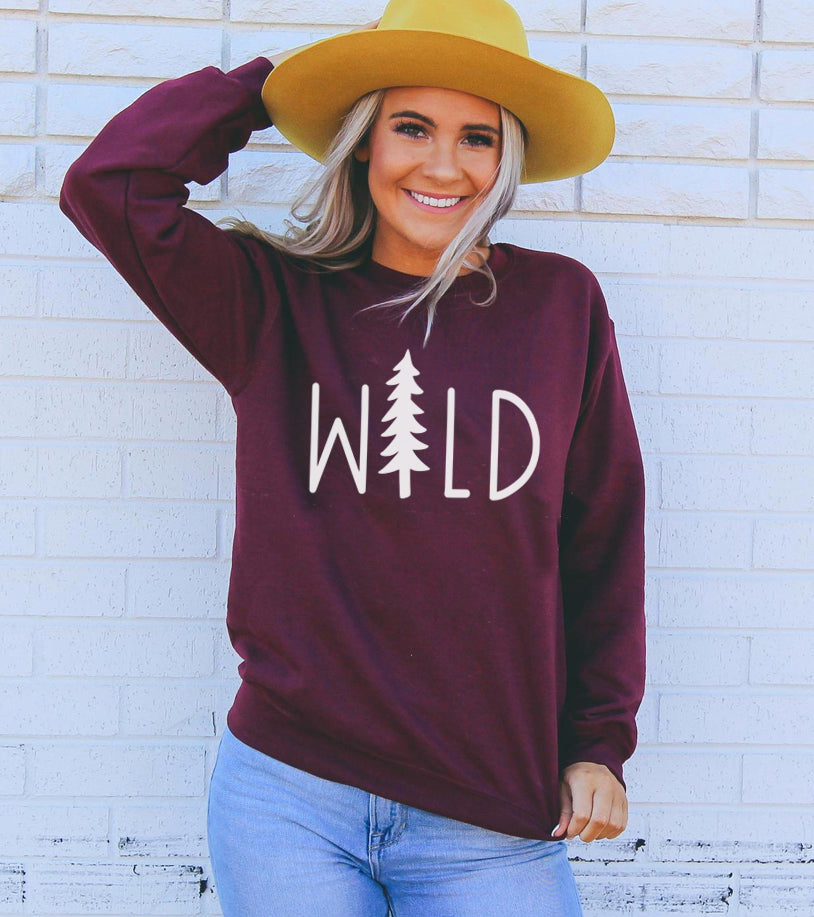 Woman in a yellow hat and maroon "WILD" sweater, posing cheerfully against a white brick wall.