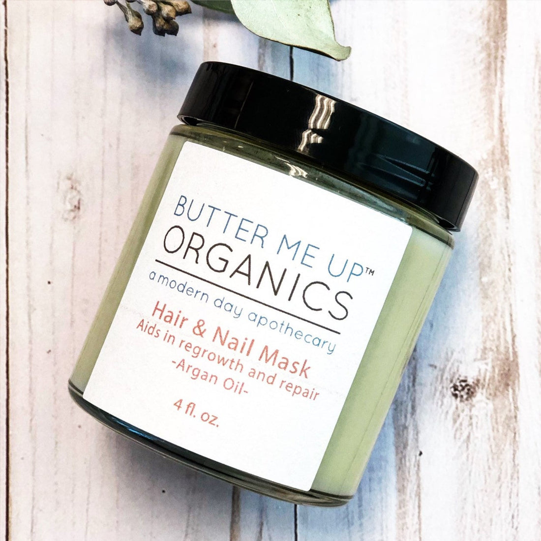 Green jar with black lid, labeled "Butter Me Up Organics Hair &amp; Nail Mask" on wood surface.