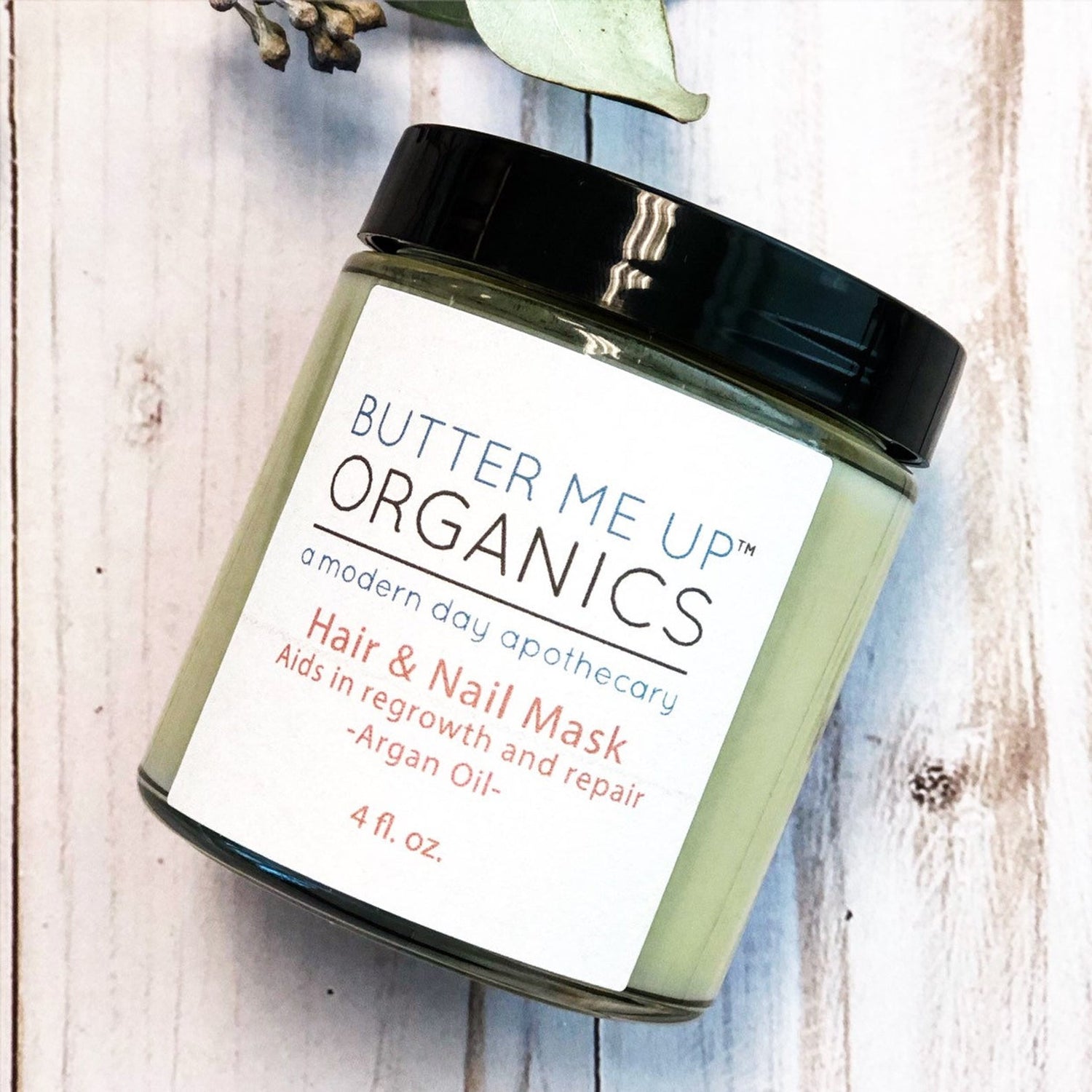 Green jar with black lid, labeled "Butter Me Up Organics Hair &amp; Nail Mask" on wood surface.