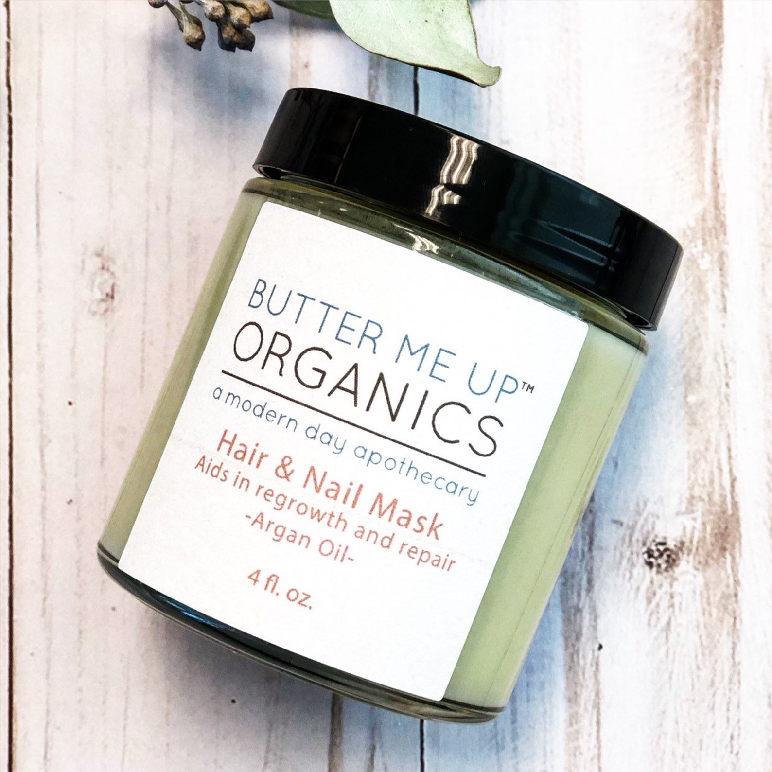 Green jar with black lid, labeled "Butter Me Up Organics Hair &amp; Nail Mask" on wood surface.