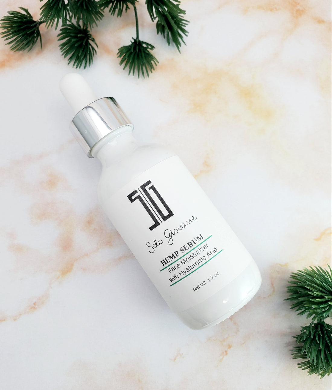 White serum bottle with dropper cap on marbled surface, surrounded by green pine sprigs.