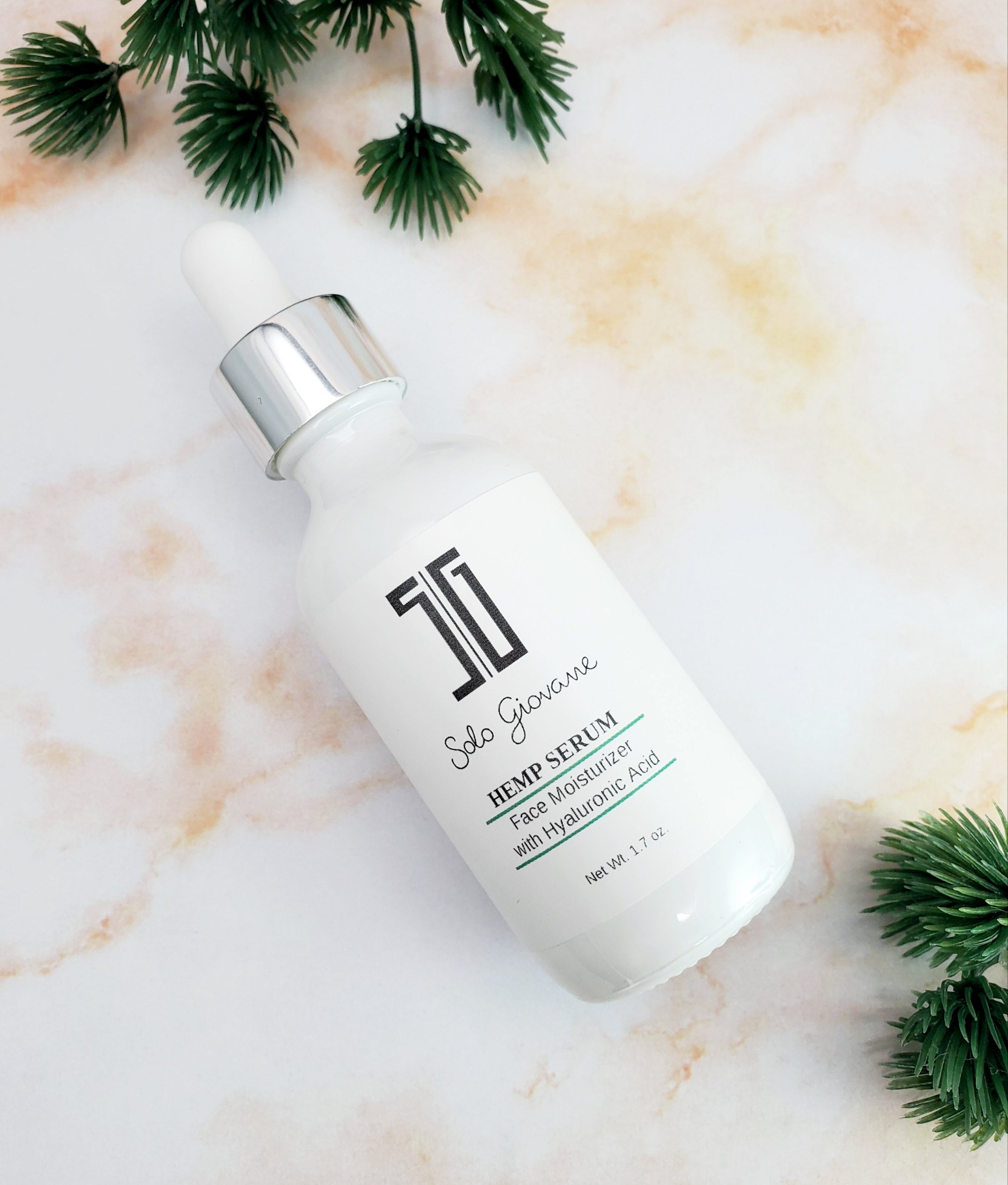 White serum bottle with dropper cap on marbled surface, surrounded by green pine sprigs.