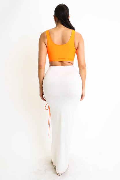 Woman in orange top and fitted white skirt, standing on white background, facing away.