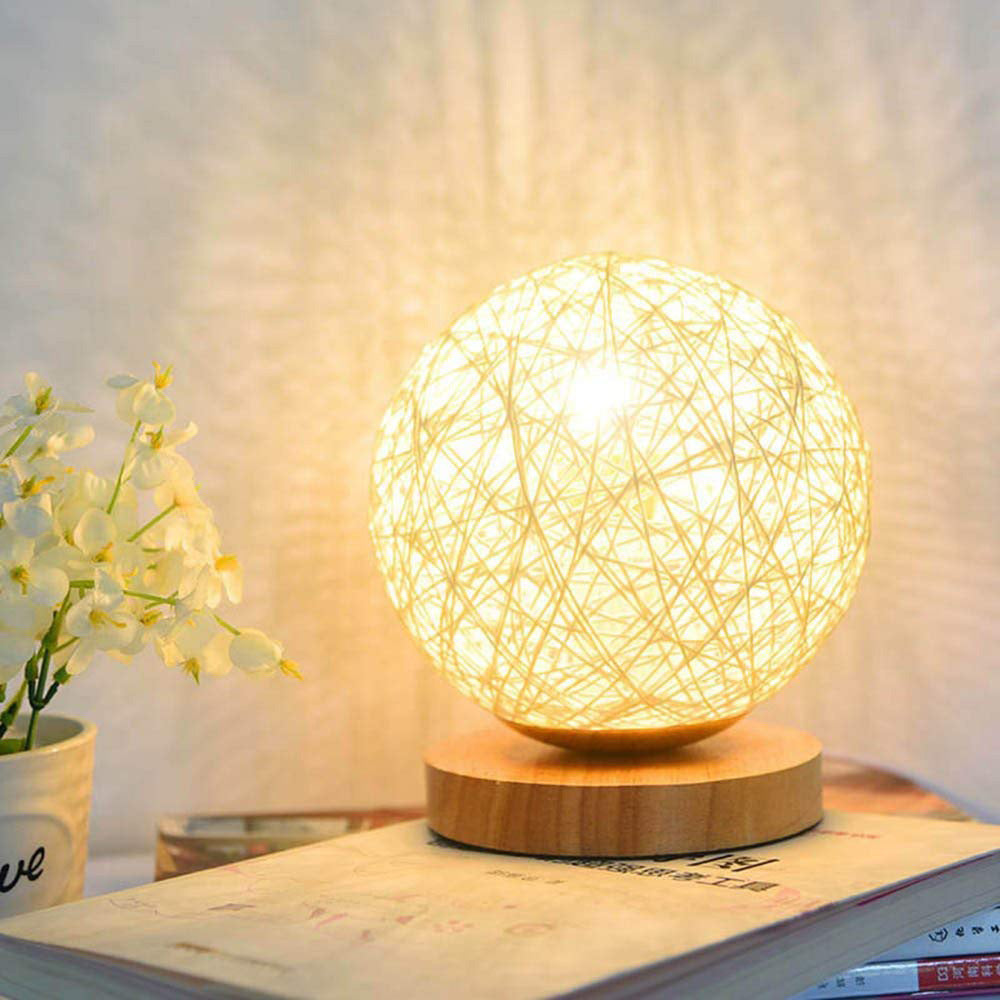 Glowing woven sphere lamp on wooden base next to a small vase of white flowers on a book.