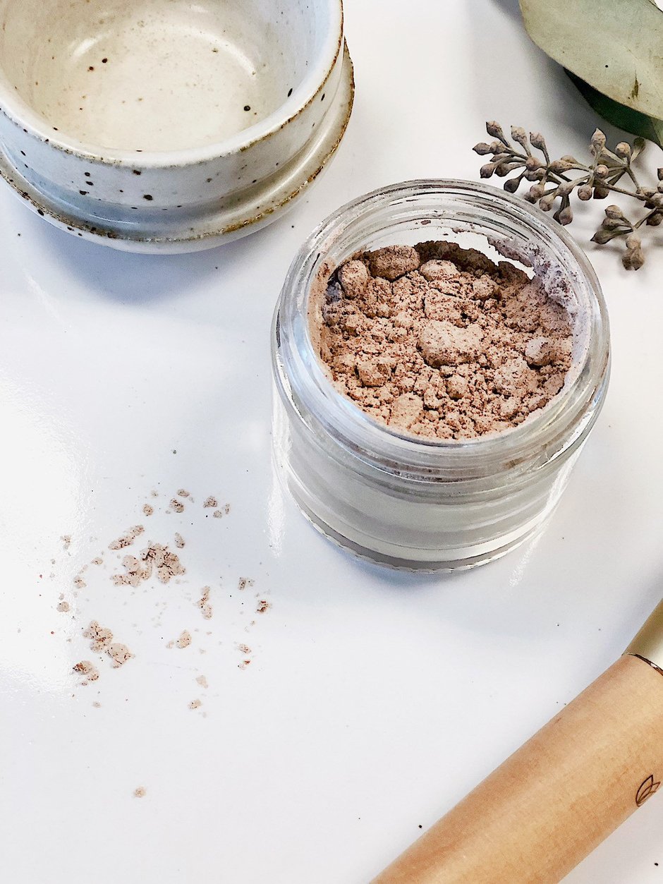 Open jar with pink powder on white surface, scattered nearby; beige brush, small dish, and leaves.