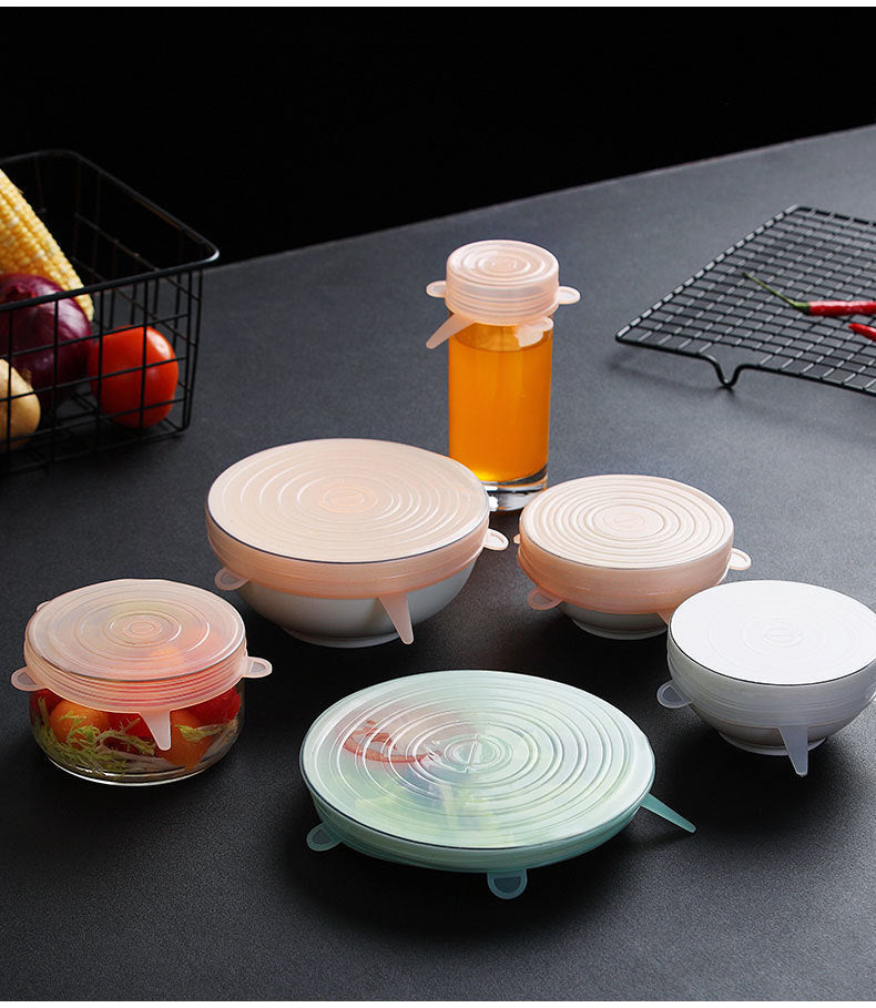 Various silicone lids on food containers and a jar on a dark surface, with fruits in the background.