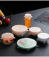 Various silicone lids on food containers and a jar on a dark surface, with fruits in the background.