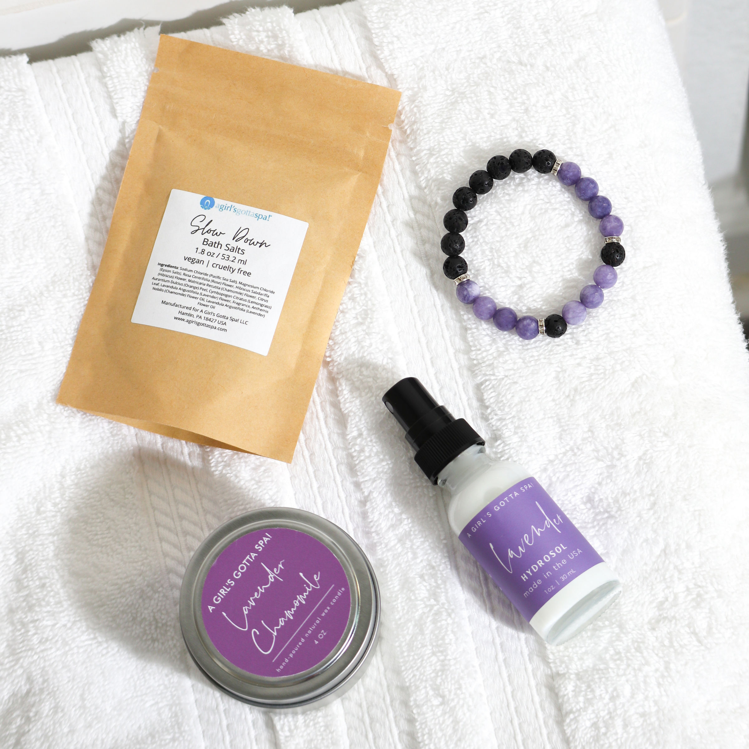 Bath salts, purple bracelet, lavender tin, and spray bottle on white towel.