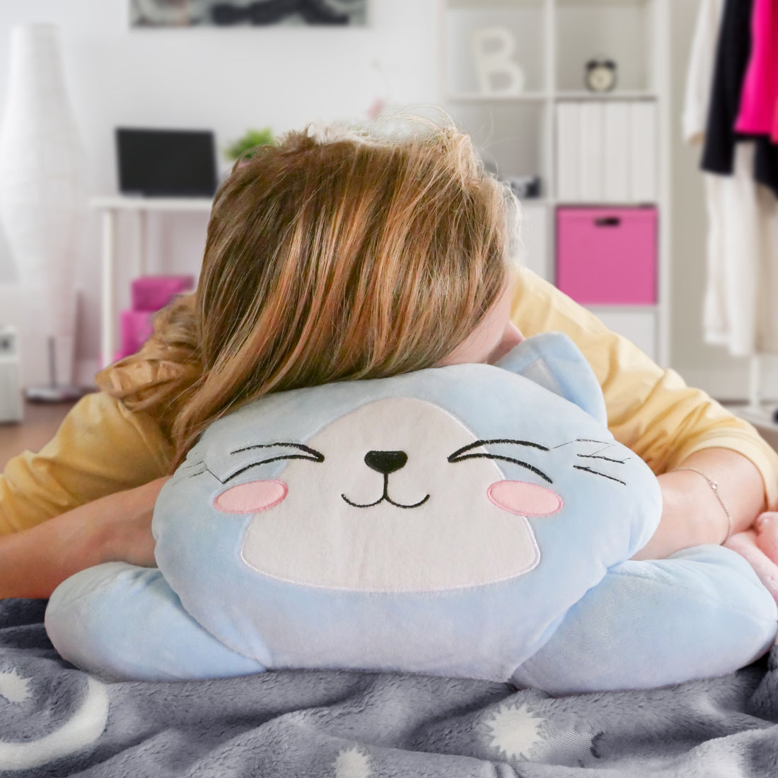Person hugging a smiling blue cat pillow with pink cheeks, in a cozy room with soft colors.