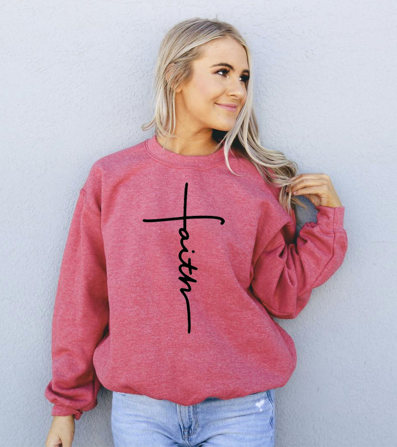 Woman in a red sweatshirt with "faith" in black, stands against a light gray wall.