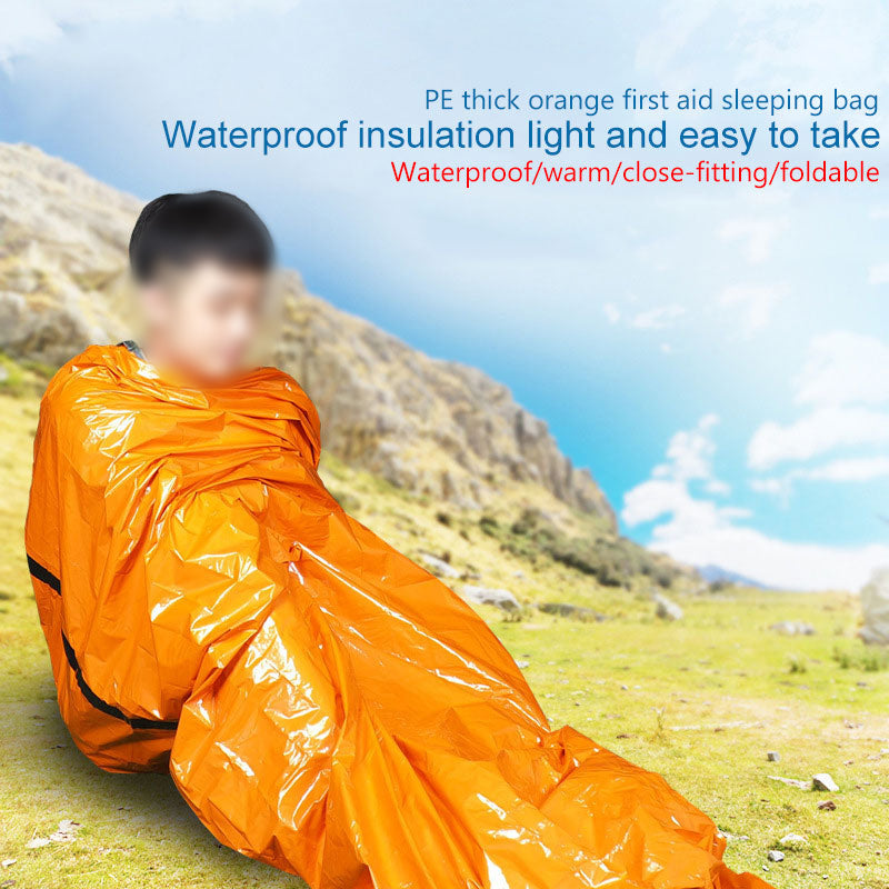 Person wrapped in shiny orange sleeping bag on grassy mountain landscape under clear blue sky.