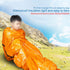 Person wrapped in shiny orange sleeping bag on grassy mountain landscape under clear blue sky.