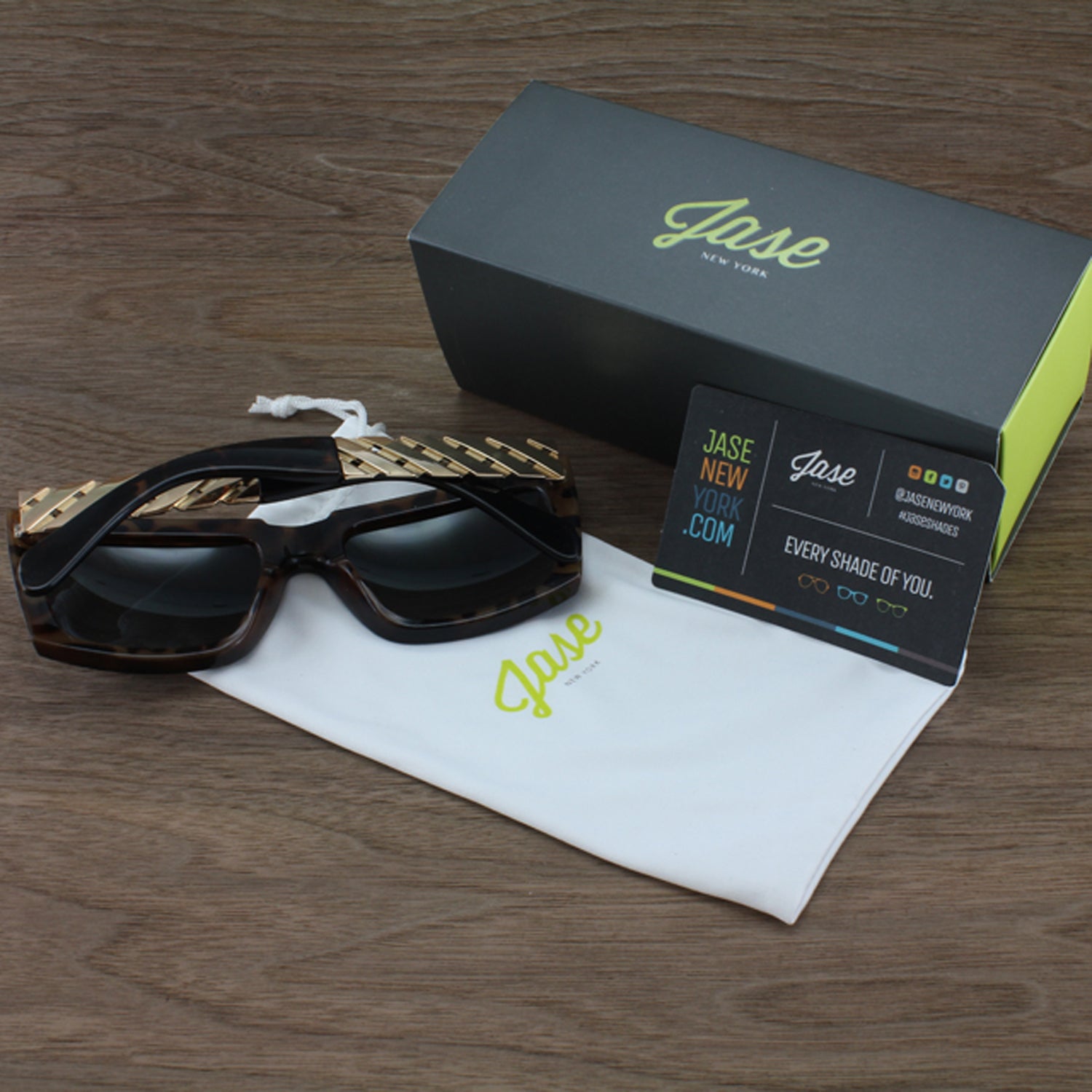 Sunglasses with gold accents, black box, white pouch, and business card on a wooden surface.