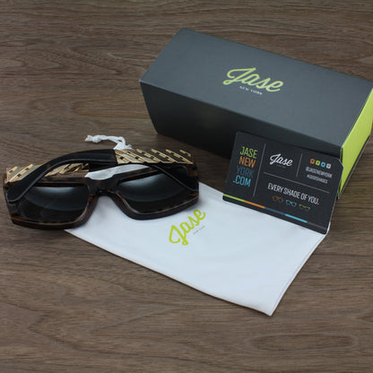 Sunglasses with gold accents, black box, white pouch, and business card on a wooden surface.