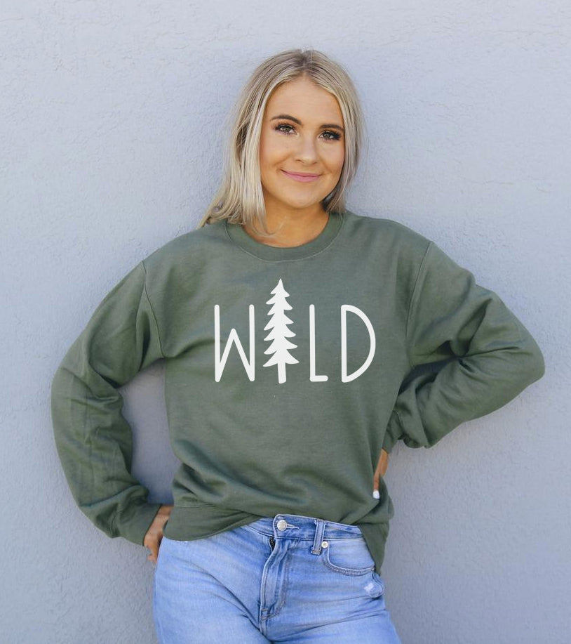 Woman in green "WILD" sweater with tree design, against a light gray wall.