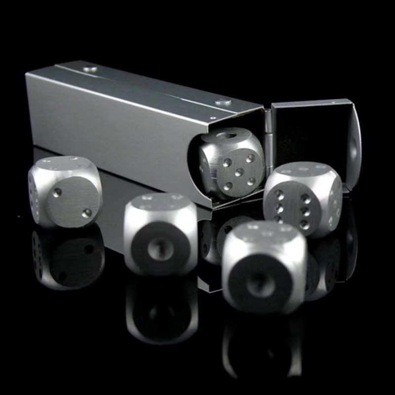 Silver metallic dice with black dots scattered near a rectangular case on a reflective black surface.