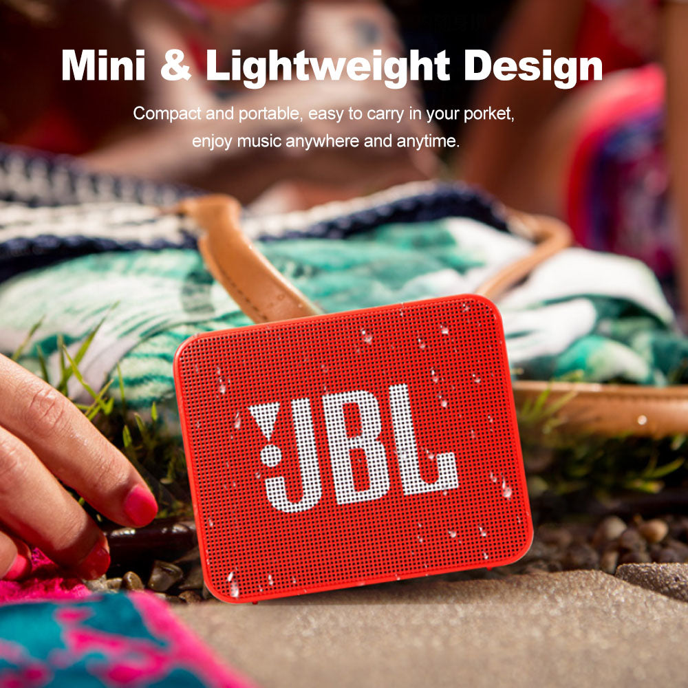 Red portable speaker with white logo, water droplets, near a colorful blanket and grass.