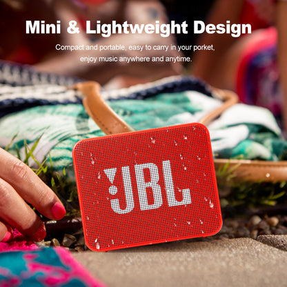Red portable speaker with white logo, water droplets, near a colorful blanket and grass.