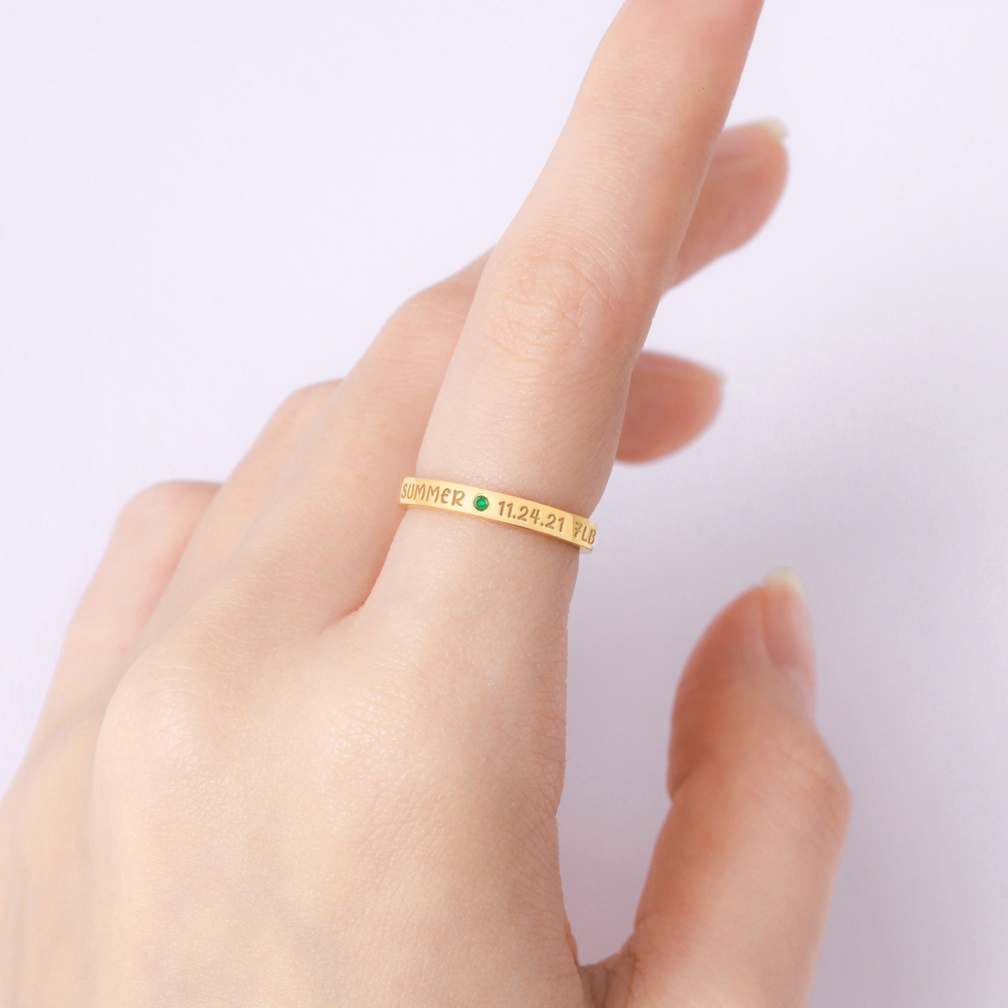 A hand with a gold ring inscribed with text and a small green gemstone, set against a pale background.