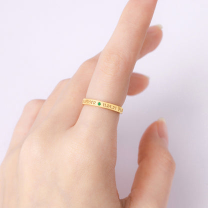 A hand with a gold ring inscribed with text and a small green gemstone, set against a pale background.