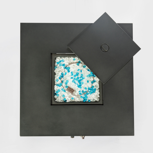 Square fire pit with a matte black surface, lid askew, showing blue and white glass beads inside.