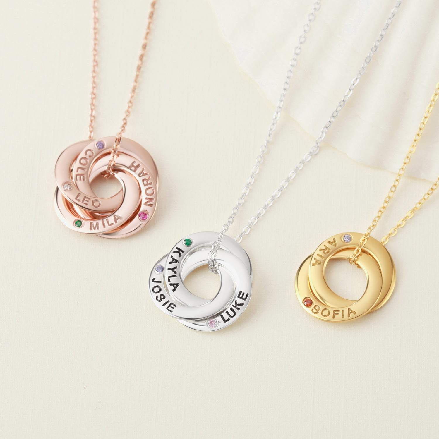 Three intertwined rings on chains: rose gold, silver, and gold, each personalized with names.