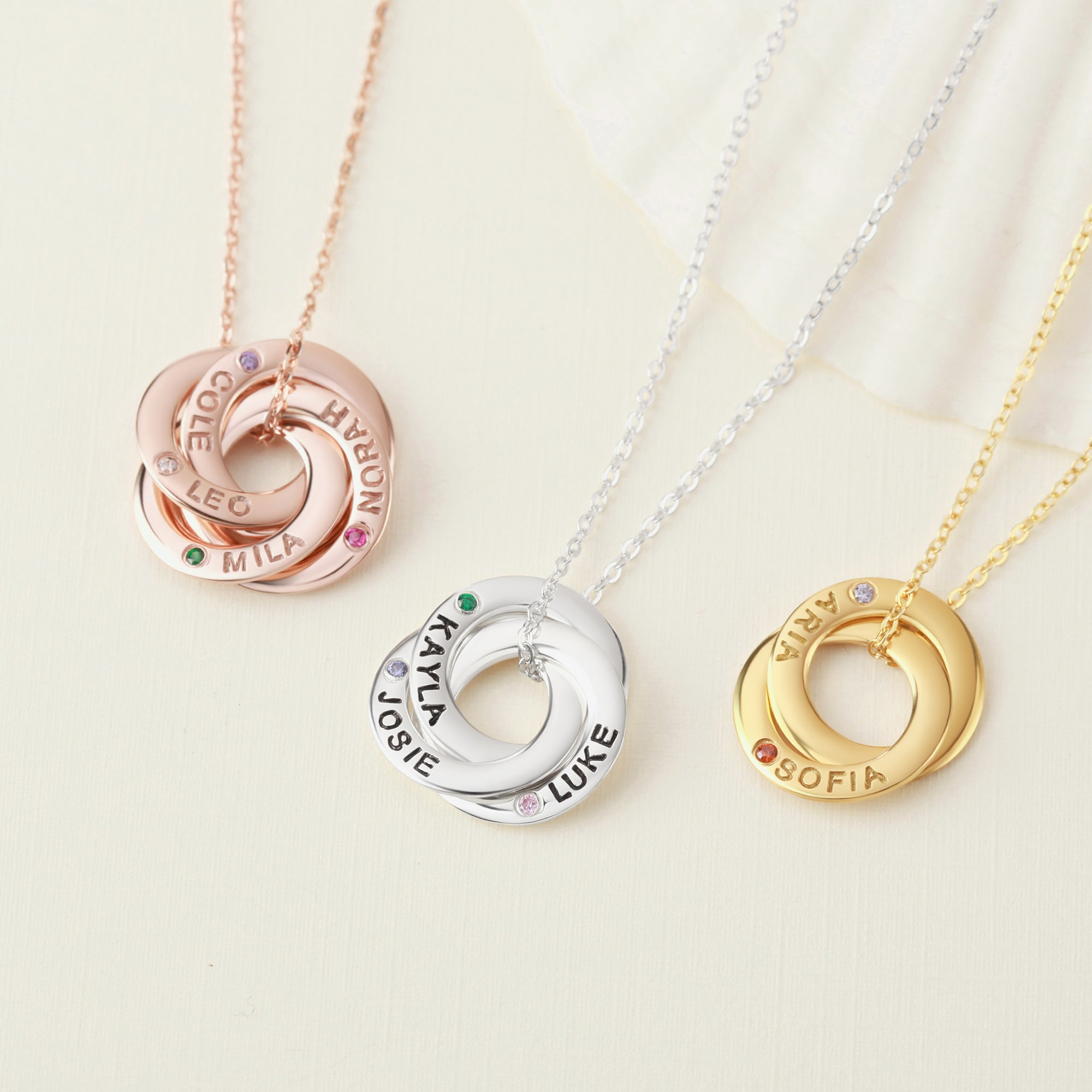Three intertwined rings on chains: rose gold, silver, and gold, each personalized with names.