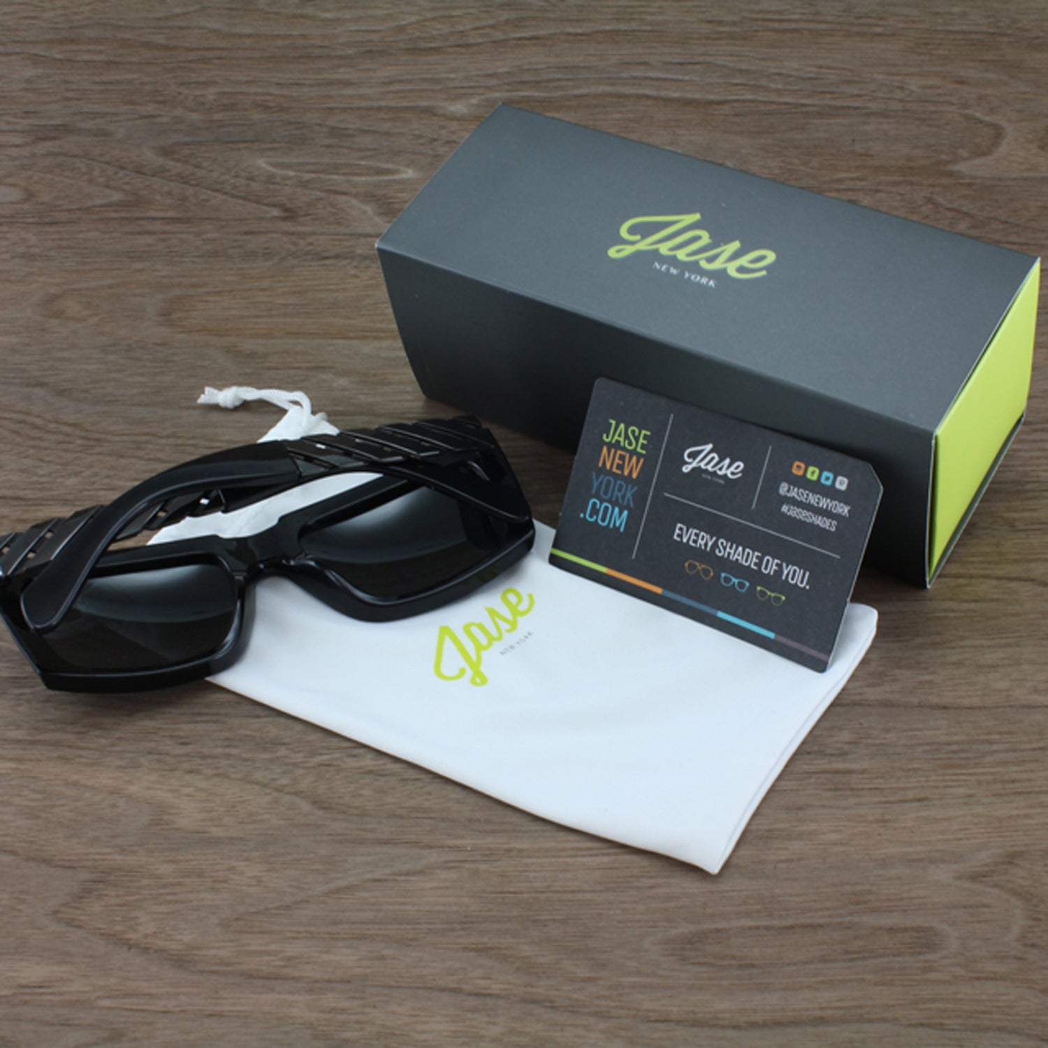 Black sunglasses, white pouch, and dark green box with lime text on a wooden surface.
