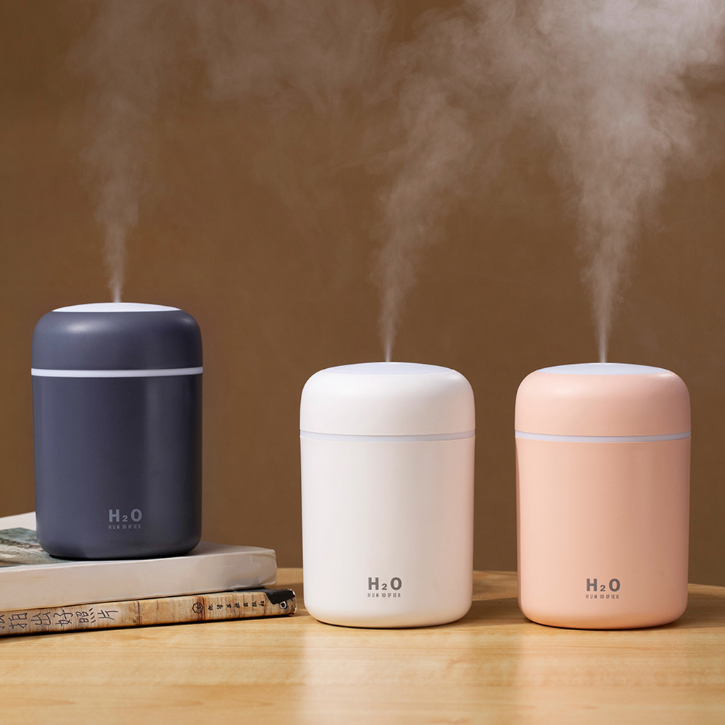 Three humidifiers emitting mist, in navy, white, and peach colors, placed on a wooden table.