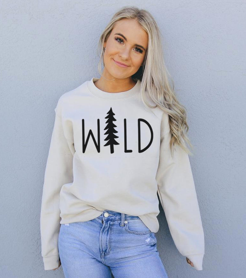Smiling person in white "WILD" sweater with tree design, wearing light blue jeans against gray wall.