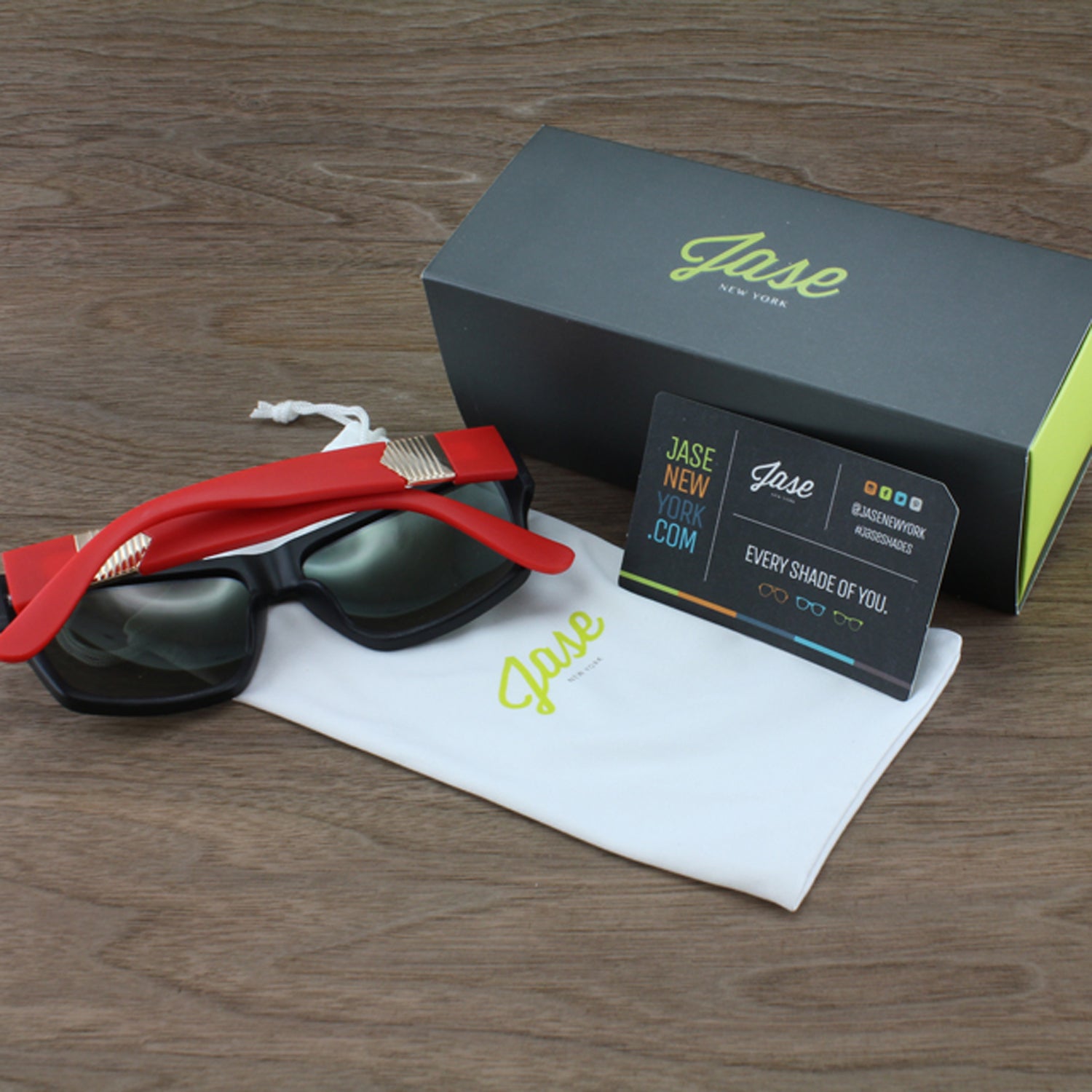 Red sunglasses, black lenses, gray box, and white pouch on wooden surface.