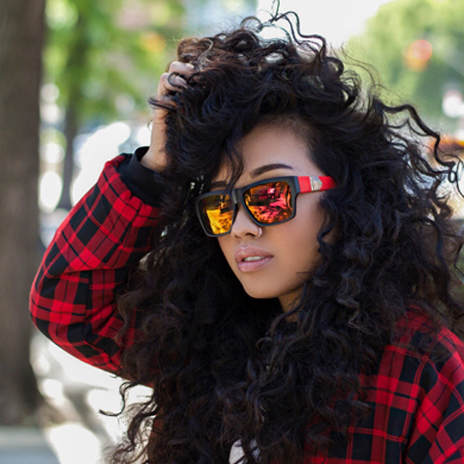 Person with curly hair in red sunglasses and a plaid shirt, outdoors with blurred trees.