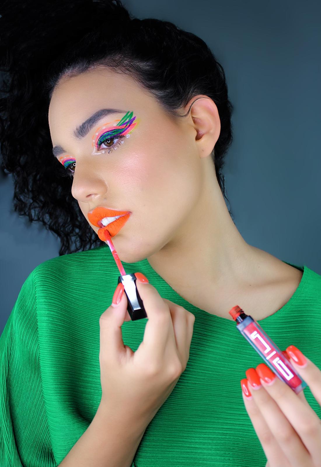 Woman with vibrant makeup and orange lipstick, colorful eyeshadow, wears green top on blue background.