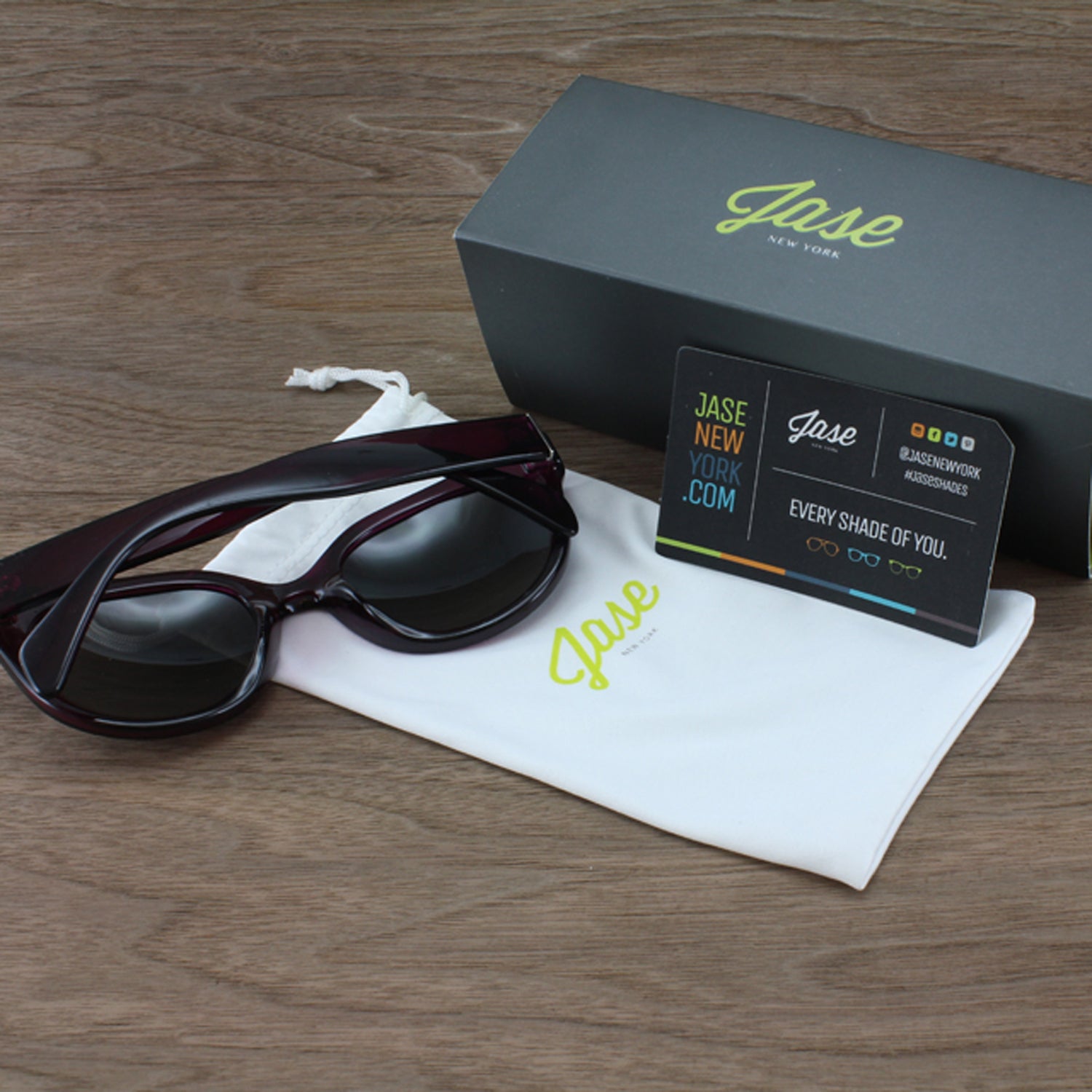 Sunglasses on a white pouch with a Jase box, business card, on a wood surface.