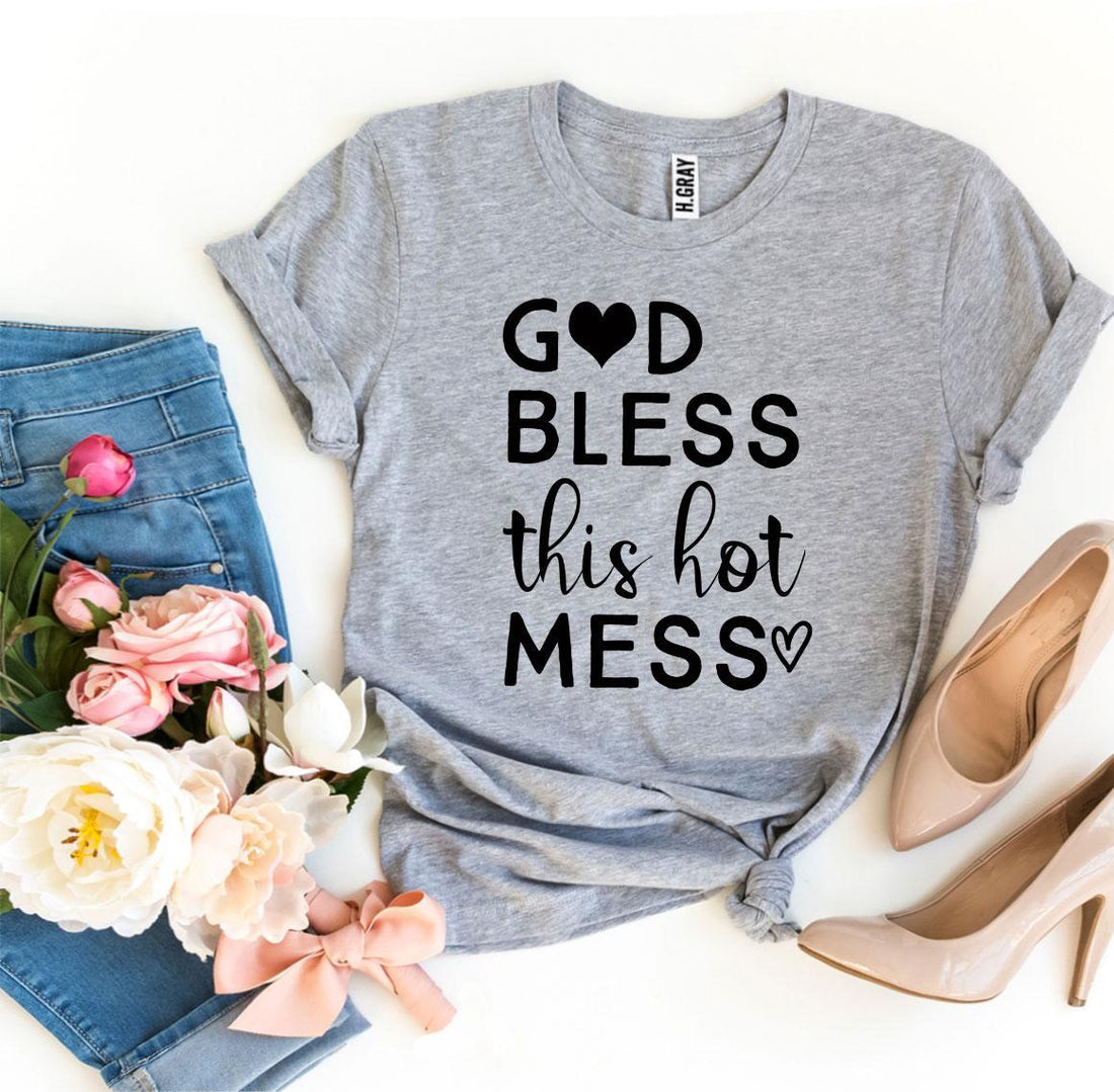 Gray t-shirt with "God bless this hot mess" text, next to pink heels, jeans, and flowers.